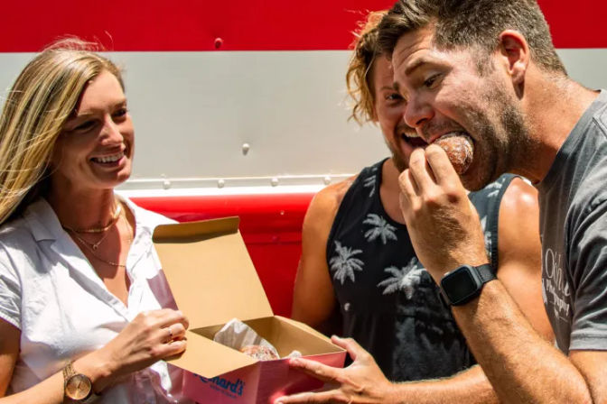 Three friends laughing by a red-and-white food truck as a man bites into a powdered sugar donut from a pink pastry box.