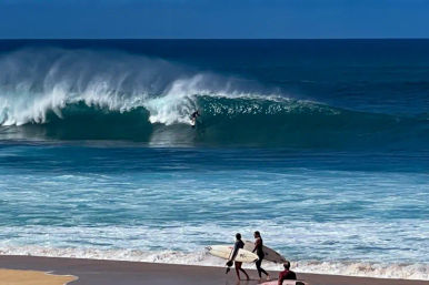 Three surfers on a sandy beach carrying boards toward a turquoise ocean while a giant blue barrel wave breaks and a surfer rides inside.