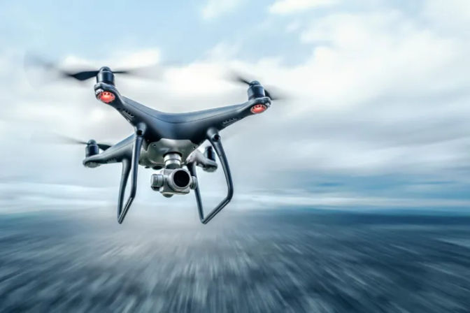 Black quadcopter drone with camera hovering over a blurred water horizon under a cloudy sky, capturing aerial footage.