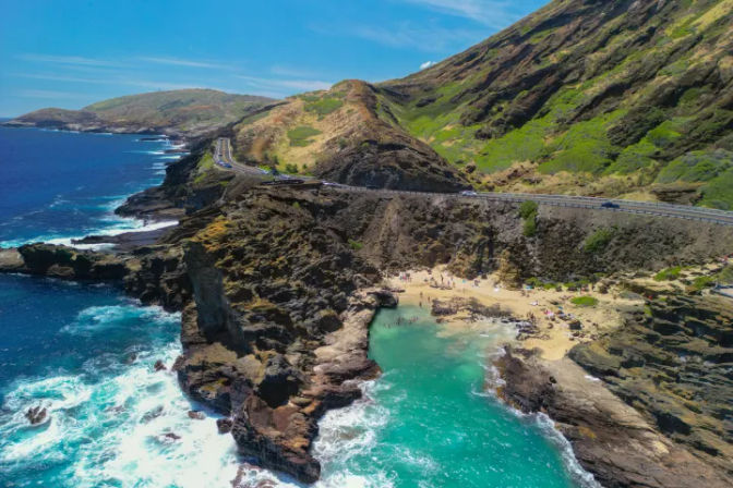 Aerial view of a scenic coastal highway winding along rugged green cliffs above a turquoise cove and tiny sandy beach with rocky outcrops and crashing ocean waves.