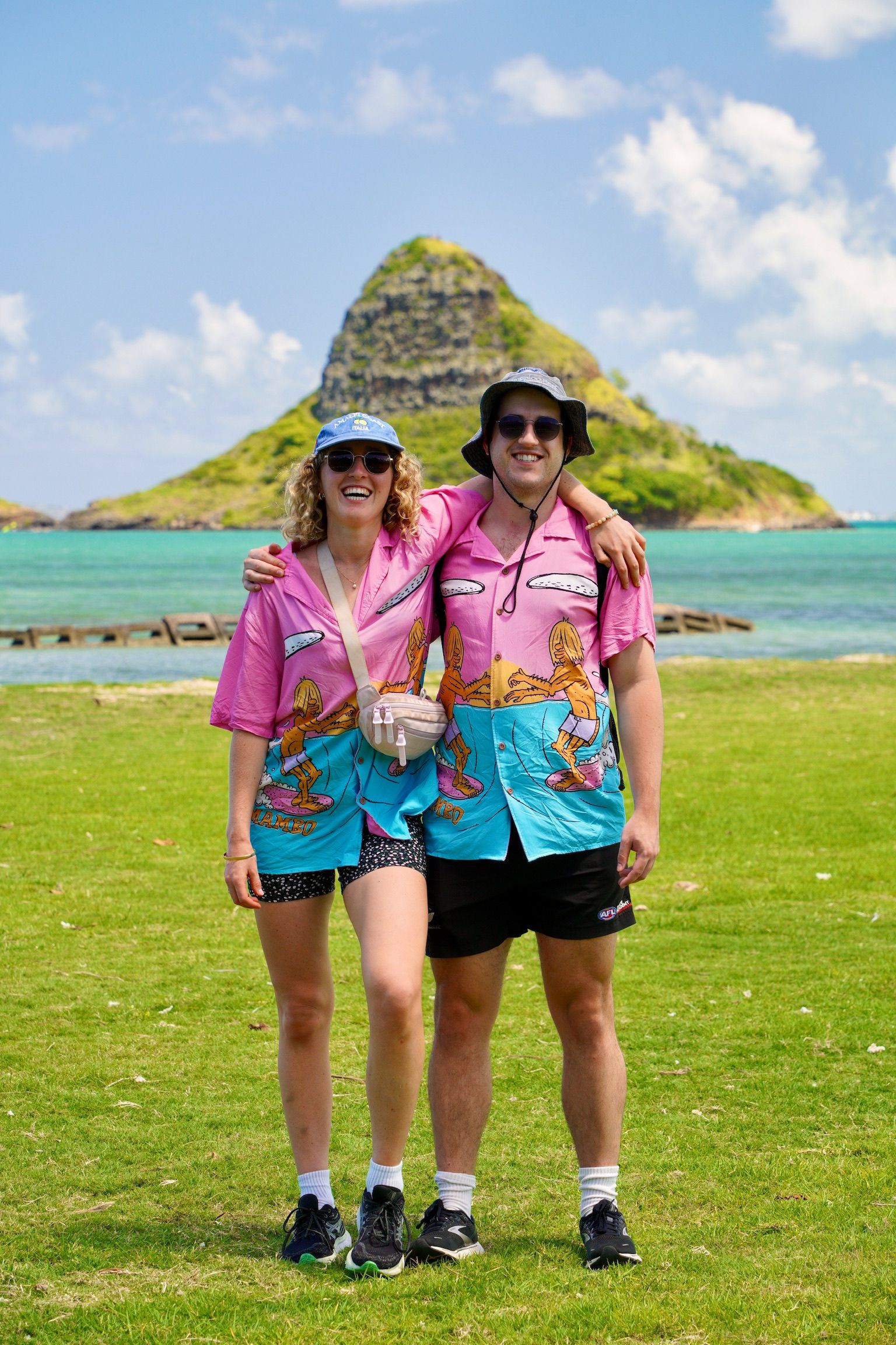 Smiling couple in matching pink-and-blue Hawaiian shirts, sunglasses and bucket hats posing arm-in-arm on a grassy shore with turquoise ocean and a small conical island under a bright blue sky.