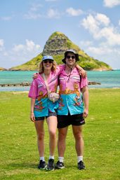 Smiling couple in matching pink-and-blue Hawaiian shirts, sunglasses and bucket hats posing arm-in-arm on a grassy shore with turquoise ocean and a small conical island under a bright blue sky.