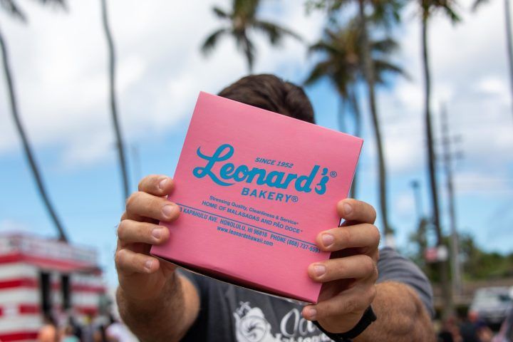 Hands presenting a pink bakery box to the camera with blurred palm trees and blue sky behind, evoking a sunny Hawaii street-food vibe.