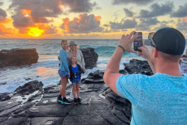 Three people posing on dark coastal rocks at a golden sunset with waves crashing, while someone in the foreground snaps a smartphone photo.