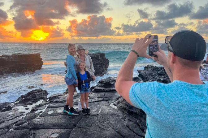 Three people posing on dark coastal rocks at a golden sunset with waves crashing, while someone in the foreground snaps a smartphone photo.