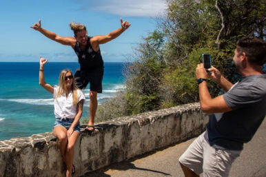Three friends at a tropical coastal overlook: woman seated on a stone wall, man striking a shaka pose on the wall, another man snapping a smartphone photo with turquoise ocean and cliffs behind them.