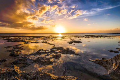 Golden sunset over rocky tidal flats and shallow tide pools reflecting dramatic clouds and a calm ocean horizon on a coastal shoreline.
