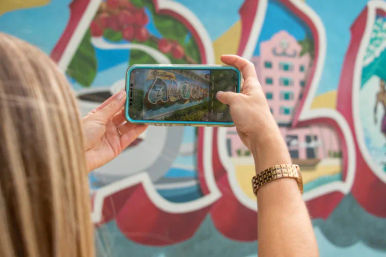 Person holding smartphone to photograph a colorful postcard-style seaside mural and street art; teal phone case, gold watch, and blonde hair visible.