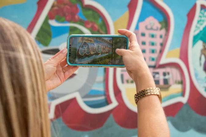 Person holding smartphone to photograph a colorful postcard-style seaside mural and street art; teal phone case, gold watch, and blonde hair visible.