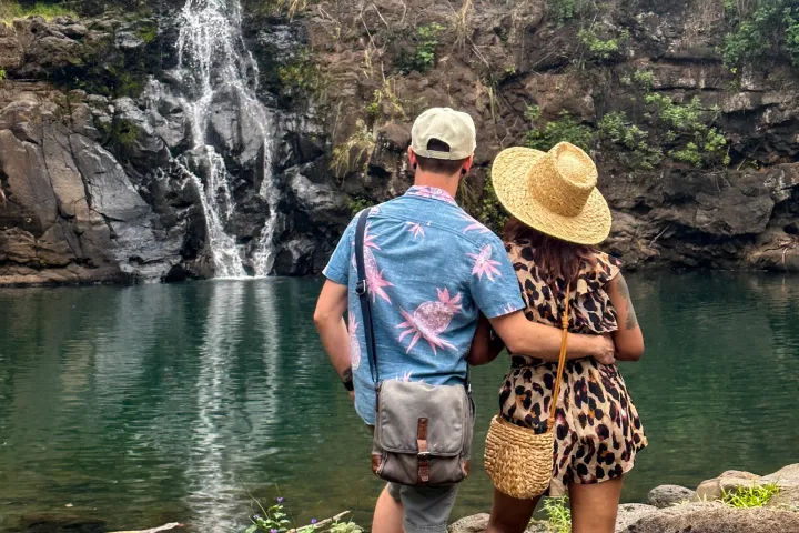 Couple from behind embracing on a rocky shore, looking at a narrow waterfall feeding a green natural pool framed by rugged cliffs; woman in a straw hat and leopard-print romper, man in a blue tropical shirt with a crossbody bag