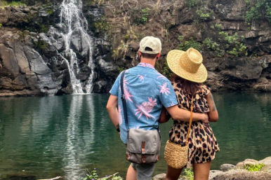 Couple from behind embracing on a rocky shore, looking at a narrow waterfall feeding a green natural pool framed by rugged cliffs; woman in a straw hat and leopard-print romper, man in a blue tropical shirt with a crossbody bag