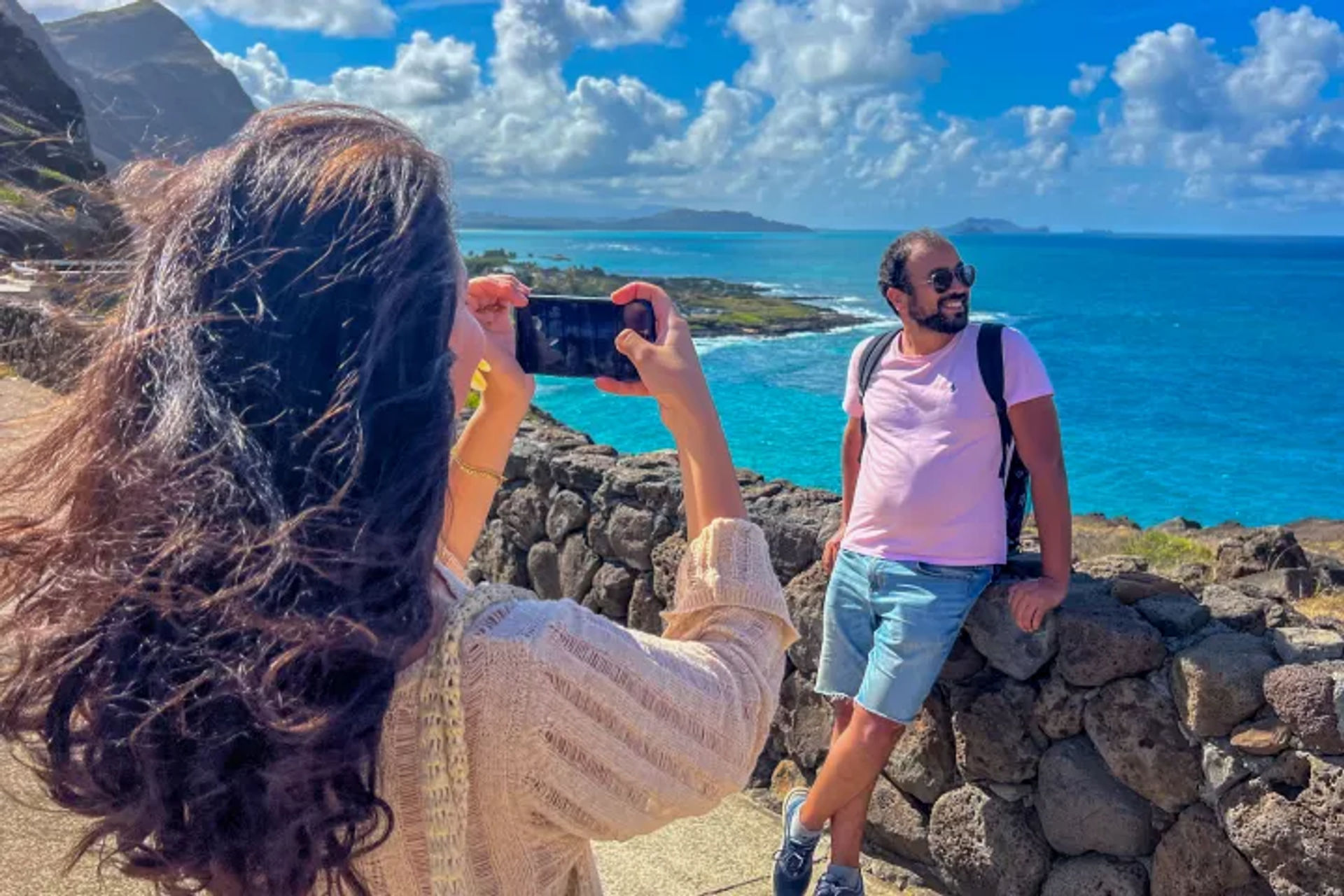 Traveler photographing a friend leaning on a stone wall with turquoise tropical ocean, rugged rocky coastline, and puffy clouds under a bright blue sky at a sunny seaside viewpoint