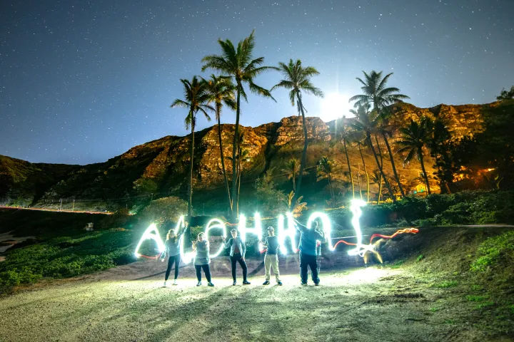 Tropical island beach at night with tall palm trees and cliffs, a group using long-exposure light painting to spell “ALOHA” beneath a starry sky.