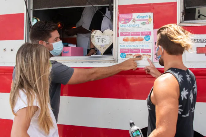 Three masked customers pointing at a food truck menu featuring malasadas at a red-and-white striped window, with a heart-shaped tip jar visible.