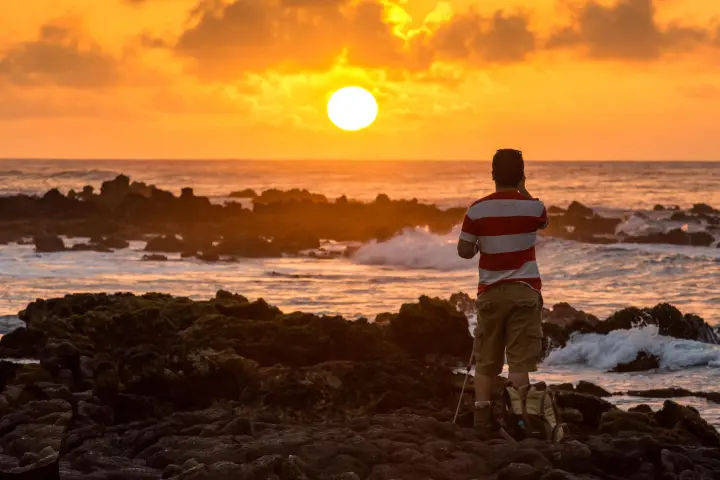 Person photographing a golden ocean sunset from a rocky shoreline with tripod and backpack nearby, waves crashing under an orange sky