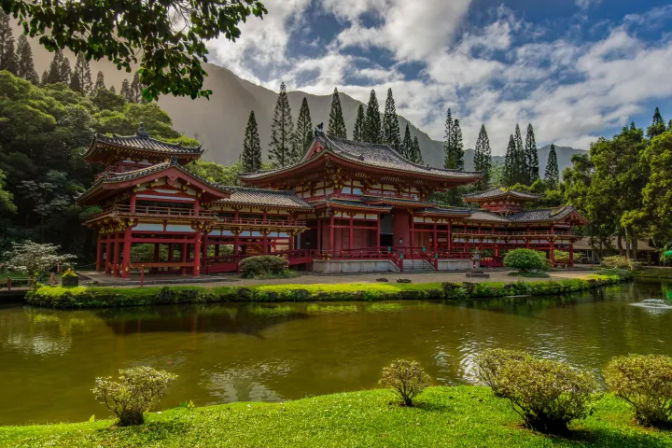 Serene red Japanese-style temple pavilion beside a reflective pond in a lush tropical mountain valley, framed by tall conifers and a dramatic cloudy sky