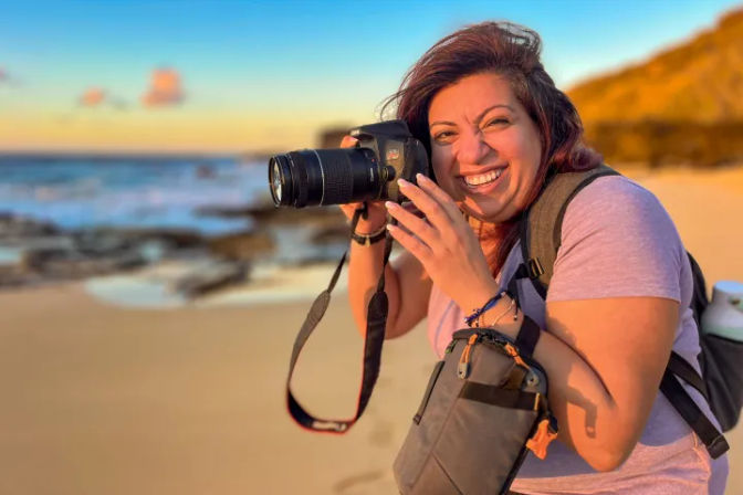 Smiling photographer holding a DSLR camera on a sandy beach during golden-hour sunset, with waves and a rocky shoreline blurred in the background.