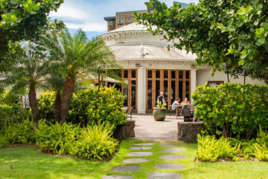 Sunny tropical garden path with stone pavers leading to a round white building with wooden doors and an outdoor cafe patio framed by palm trees and lush green shrubs.