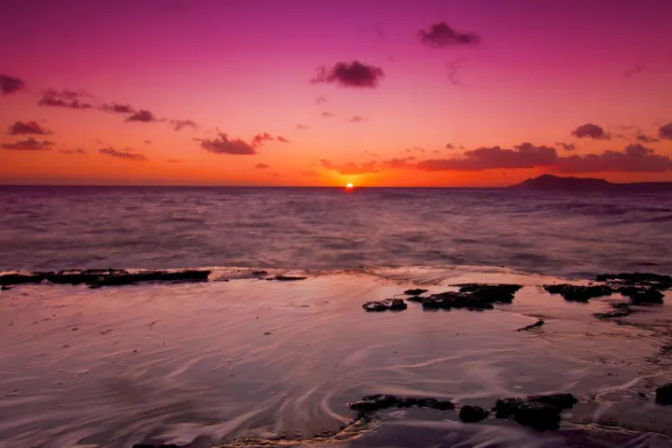 Vibrant pink and orange sunset over the ocean, rocky shoreline and wet sand reflecting the colorful sky with a small island silhouette on the horizon.