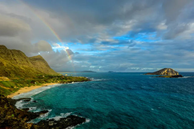 Vivid rainbow arching over rugged coastal cliffs and a sandy bay, turquoise ocean stretching to a rocky islet beneath a dramatic cloudy sky.