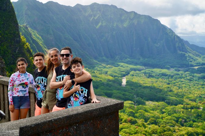Smiling family of five posing at a concrete scenic lookout above a lush tropical valley with dramatic green mountain ridges and a winding road below