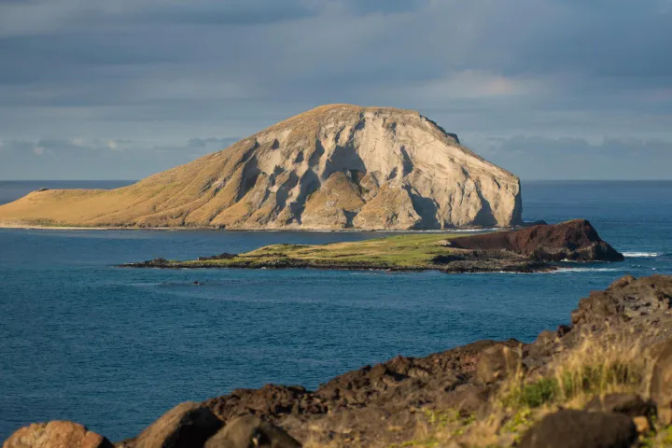 Sunlit rounded volcanic island with pale steep cliffs and grassy slopes, a smaller green-topped rocky islet in front, set in deep blue ocean under a cloudy sky, viewed from a rocky coastline.