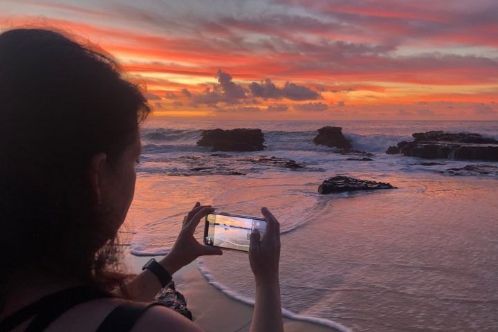 Silhouette of a person snapping a vibrant pink-orange coastal sunset over rocky shoreline with a smartphone, waves lapping wet sand that mirrors the colorful sky.