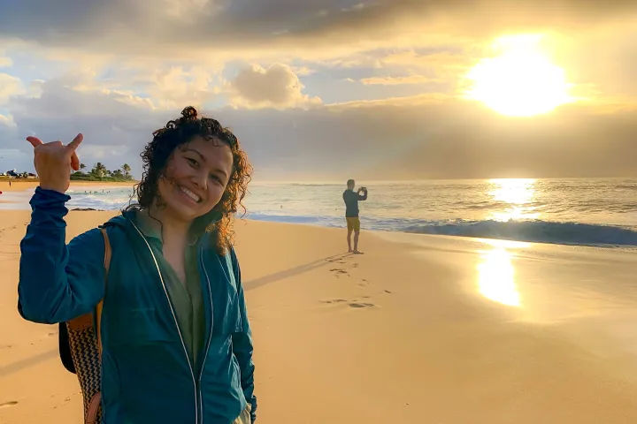 Smiling person flashing a shaka sign on a golden-sand beach at sunrise, wearing a teal jacket and backpack, with another person photographing the ocean as sunlight glints on calm waves and wet shore.