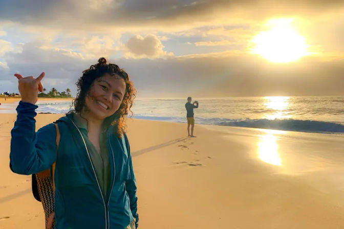 Smiling person flashing a shaka sign on a golden-sand beach at sunrise, wearing a teal jacket and backpack, with another person photographing the ocean as sunlight glints on calm waves and wet shore.