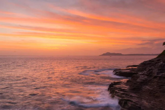 Vibrant orange-pink sunset over the ocean, waves lapping a rocky shoreline with a distant island silhouette on the horizon