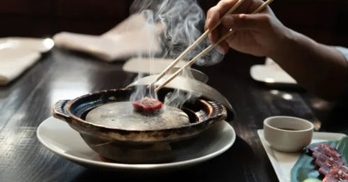 Steaming tabletop grill with chopsticks searing a thin slice of beef, dipping sauce and extra raw slices on a plate at a cozy Korean BBQ-style table.