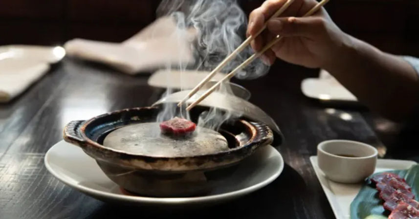 Steaming tabletop grill with chopsticks searing a thin slice of beef, dipping sauce and extra raw slices on a plate at a cozy Korean BBQ-style table.