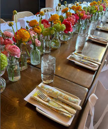 Indoor long wooden dining table set for a spring event, lined with small glass vases of colorful ranunculus and green blooms, patterned napkins, silver cutlery and tall water glasses.