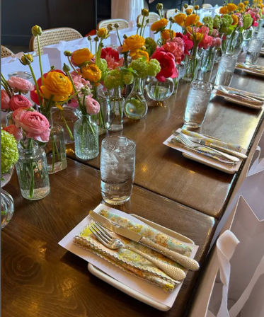 Indoor long wooden dining table set for a spring event, lined with small glass vases of colorful ranunculus and green blooms, patterned napkins, silver cutlery and tall water glasses.