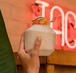 Hand holding a fresh young coconut cocktail topped with orange and watermelon garnish on a bamboo skewer, pink neon sign and tropical bar backdrop