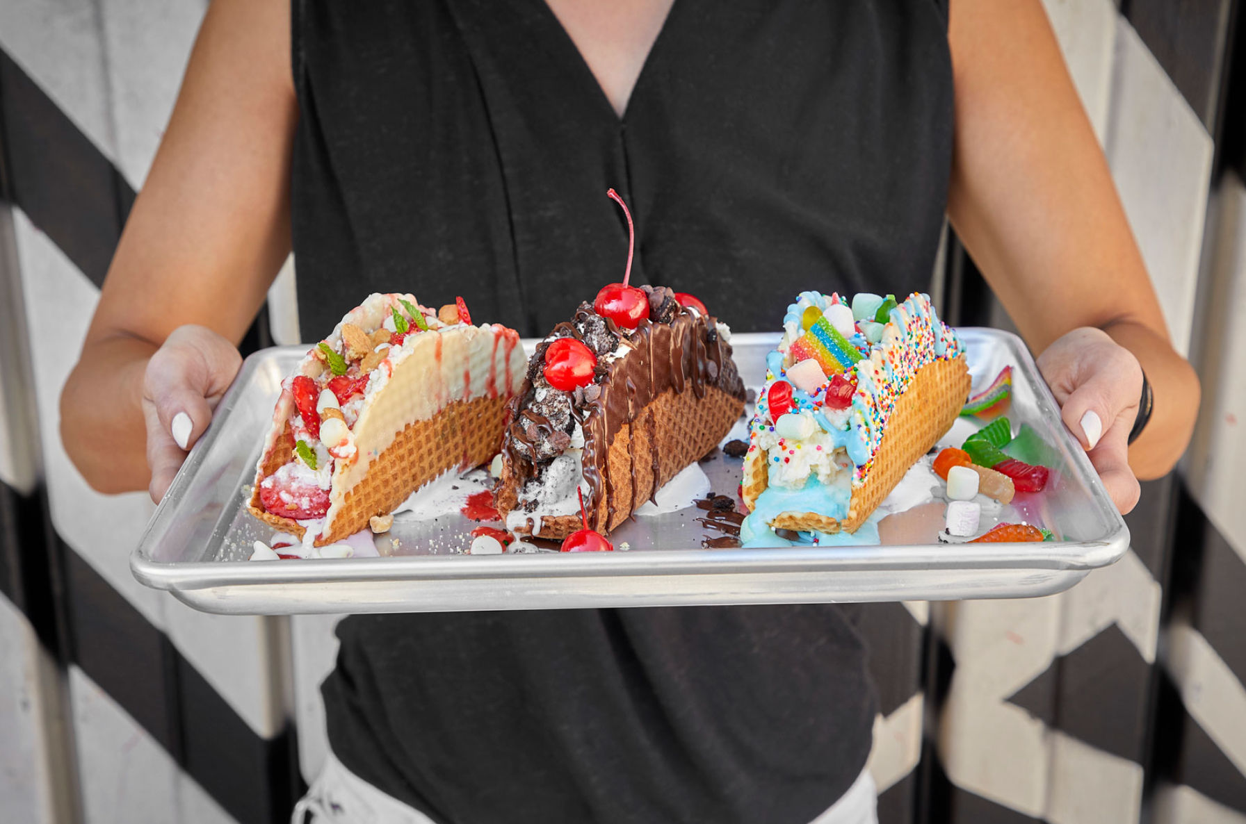 Hands hold a metal tray with three colorful waffle ice-cream tacos topped with chocolate sauce and cherries, whipped cream, sprinkles and gummy candies.