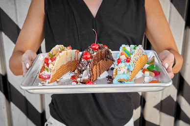 Hands hold a metal tray with three colorful waffle ice-cream tacos topped with chocolate sauce and cherries, whipped cream, sprinkles and gummy candies.