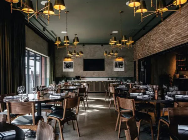 Urban upscale restaurant interior — industrial-chic dining room with exposed brick, brass pendant lights, wooden tables and chairs, large windows, and neatly set tables with wine glasses and napkins.