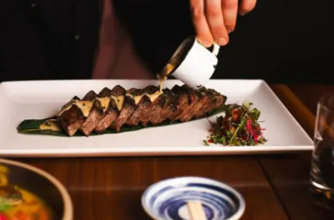 Sliced seared steak on a banana leaf with creamy sauce being poured from a small cup and microgreens garnish on a white rectangular plate at a restaurant table.
