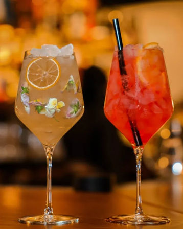 Two vibrant cocktails on a bar counter: a pale citrus spritz with ice, lemon slice and edible flower garnishes, and a bright red iced cocktail with a black straw.