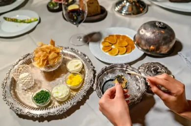 Fine-dining caviar service: hands spooning black caviar onto a blini over ornate silver dishes with crackers, small bowls of chopped egg yolk and white, chives, crème fraîche, thin toasted blinis and a wine glass on a white tablecloth.