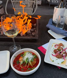Al fresco patio dining with a tabletop fire pit, a wine glass and two small plates: a fried croquette in tomato sauce topped with greens and cheese, and fritters on herbed yogurt with pickled onions.