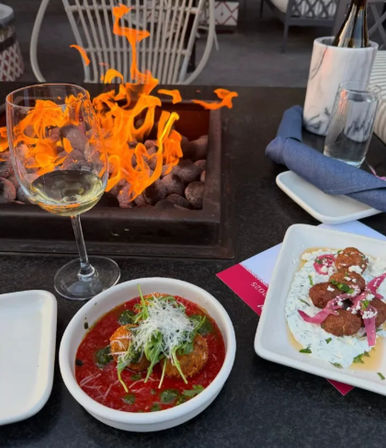Al fresco patio dining with a tabletop fire pit, a wine glass and two small plates: a fried croquette in tomato sauce topped with greens and cheese, and fritters on herbed yogurt with pickled onions.