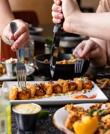Crispy fried shrimp skewer on a rectangular plate with dipping sauce and lemon wedge, hands using fork and knife at a busy seafood restaurant table — shared appetizer.