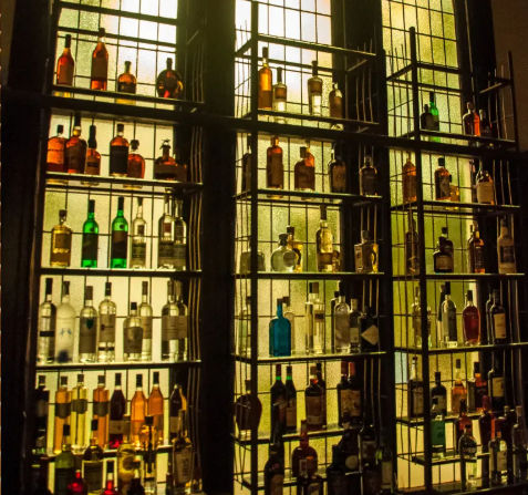 Warm backlit display of assorted liquor bottles on metal shelves against tall stained-glass windows in a cozy bar setting.