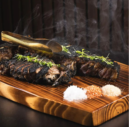 Smoky charred ribeye slices on a wooden cutting board, garnished with fresh thyme, gold tongs and three seasoning piles — wood-fired steakhouse style