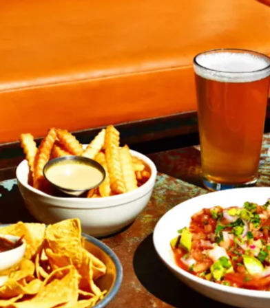 Pint of amber beer with crinkle-cut fries and dipping sauce, tortilla chips and fresh pico de gallo on a rustic bar table in a casual pub.