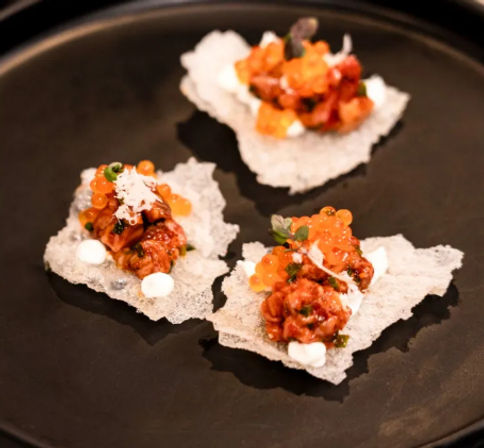 Three crispy rice cracker sushi bites topped with spicy tuna tartare, bright orange salmon roe, microgreens and dollops of creamy sauce on a matte black plate — Japanese-style gourmet appetizer.