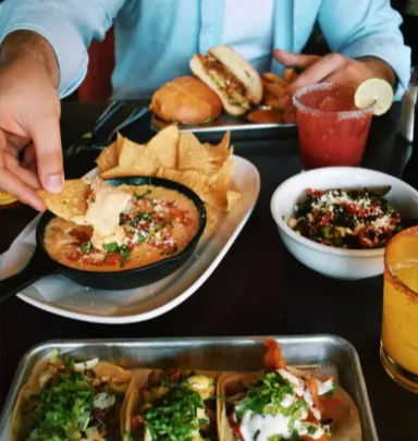 Hands reaching for a tortilla chip dipped into a sizzling skillet of queso with salsa and crema, street tacos on a metal tray, a burger, a salsa bowl and a lime-rimmed margarita on a restaurant table