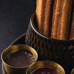 Crispy cinnamon-sugar churros standing in a textured bowl with small brass cups of chocolate and berry dipping sauces on a dark background
